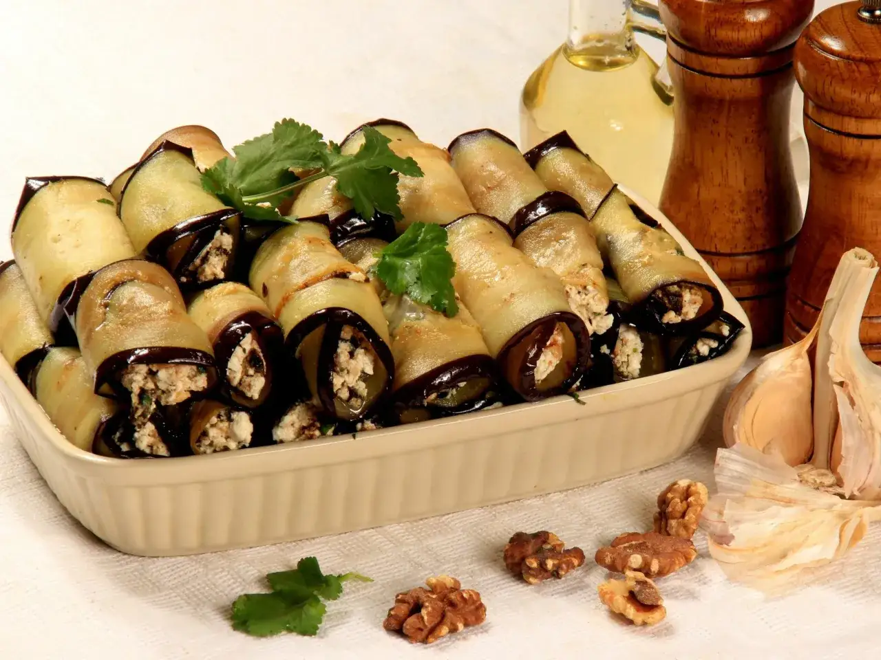 Roladki z bakłażana z nadzieniem serowym, ozdobione natką pietruszki, obok orzechy włoskie i czosnek.