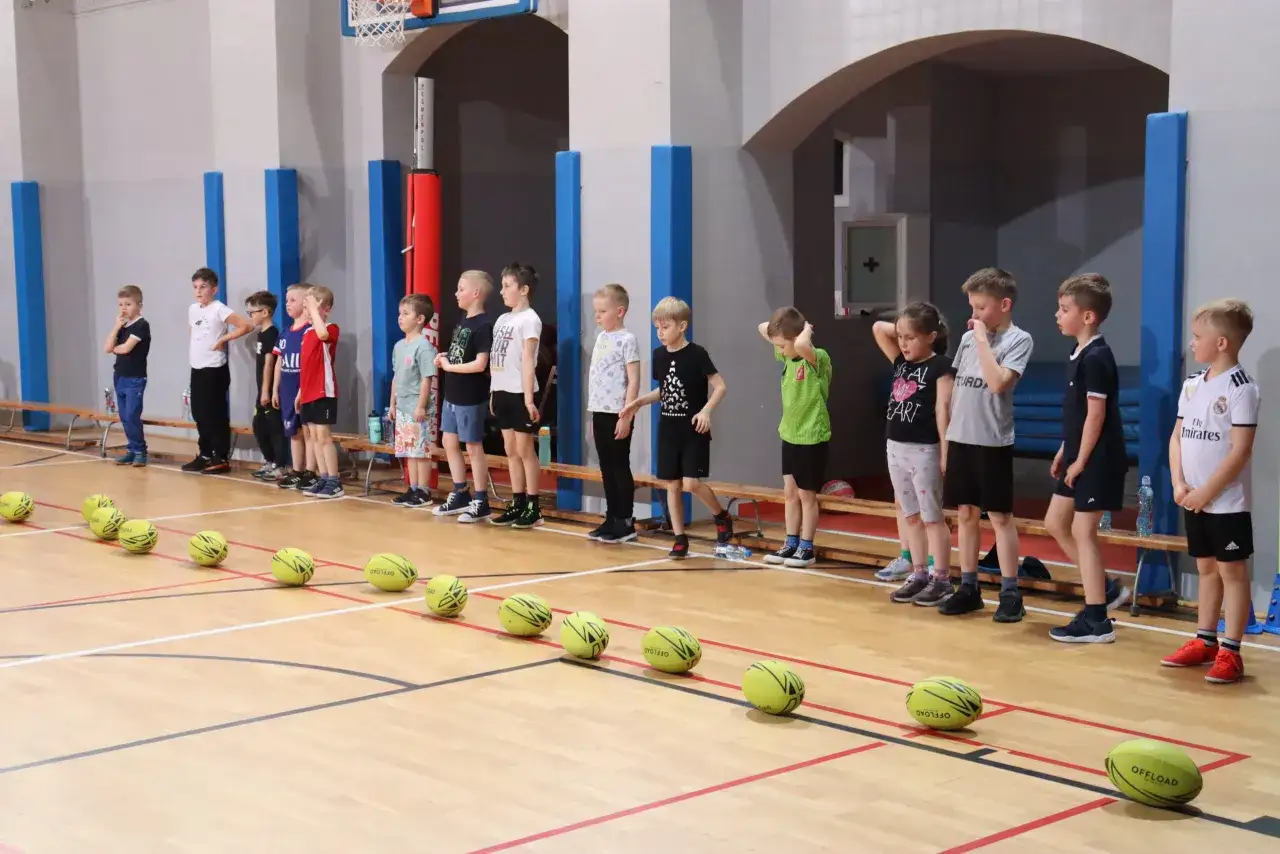 Dzieci w hali sportowej czekają na rozpoczęcie gry i zabawy. Na parkiecie leżą żółte piłki.
