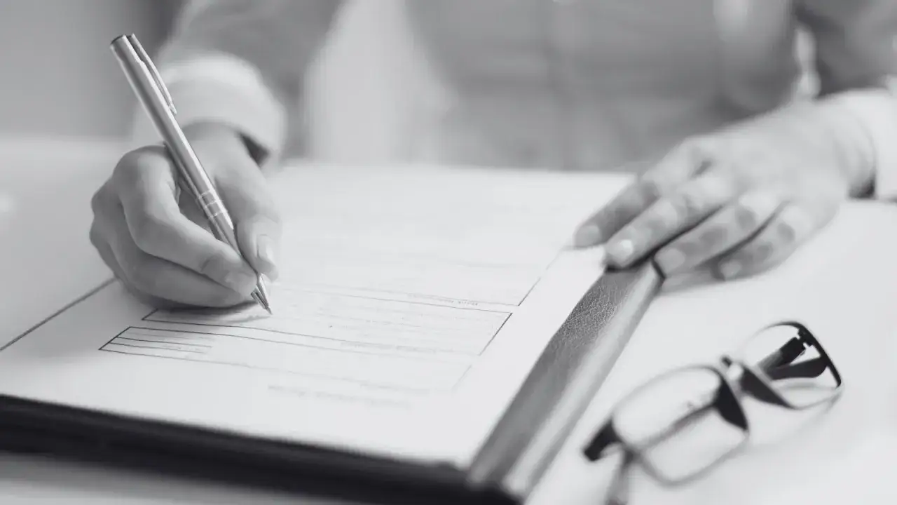 Dłoń pisze na dokumencie, obok okulary. Dowiedz się, jak sprawdzić pozwolenie na budowę sąsiada, analizując formalności.