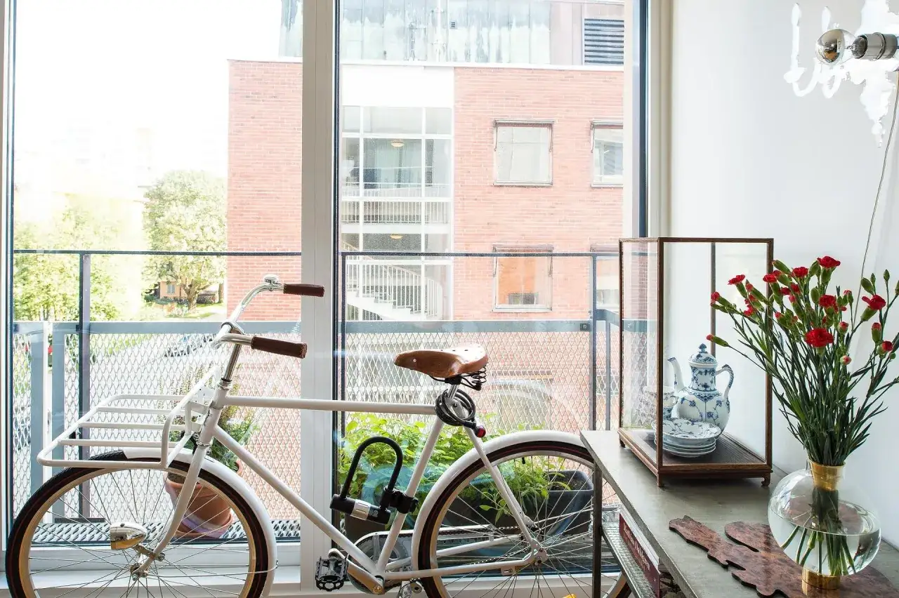 Biały rower z brązowym siodełkiem i uchwytami stoi na balkonie. Za nim widać ceglany budynek i zieleń.