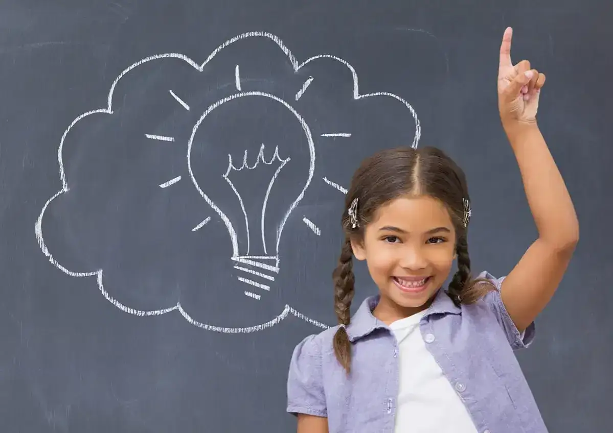 Uśmiechnięta dziewczynka z uniesionym palcem na tle tablicy z rysunkiem żarówki.