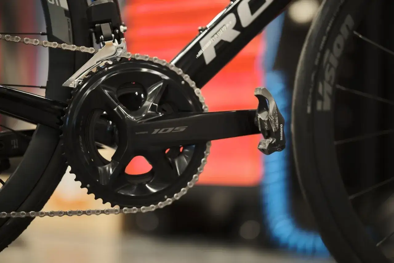 Szukasz informacji, jak dobrać korbę do roweru? Zobacz zbliżenie na czarną korbę Shimano 105 z łańcuchem i pedałem.