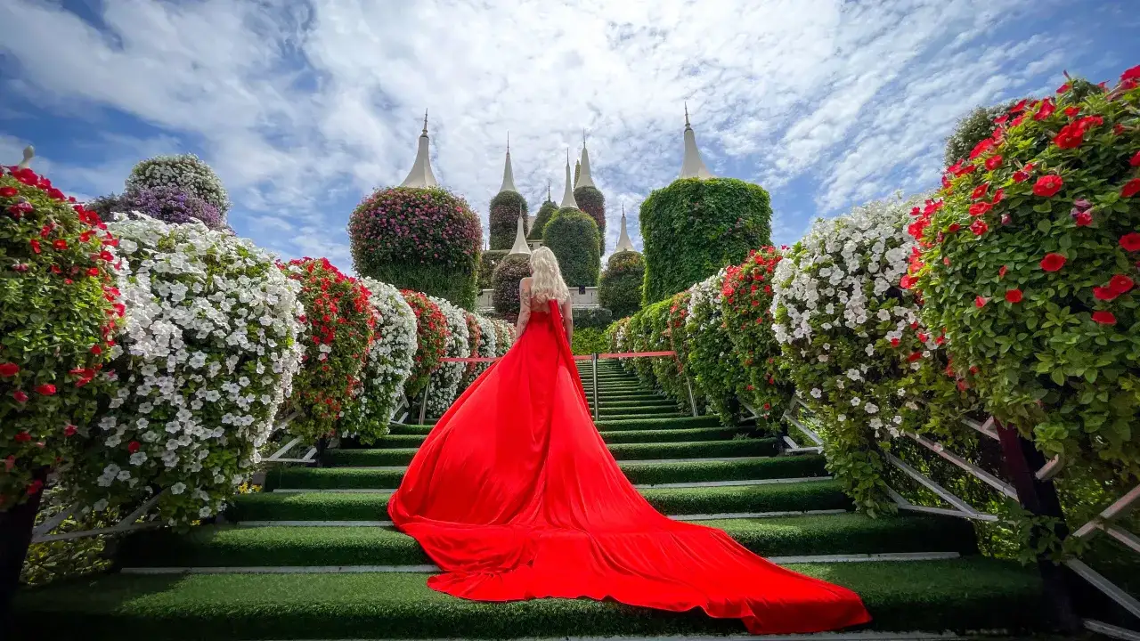 Kobieta w czerwonej sukni na schodach w Miracle Garden w Dubaju, otoczona kwitnącymi krzewami i wieżami z kwiatów.