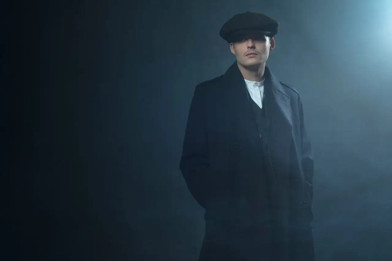 Młody mężczyzna w czapce i płaszczu, z charakterystyczną fryzurą w stylu Peaky Blinders, stoi w zadymionym mroku.