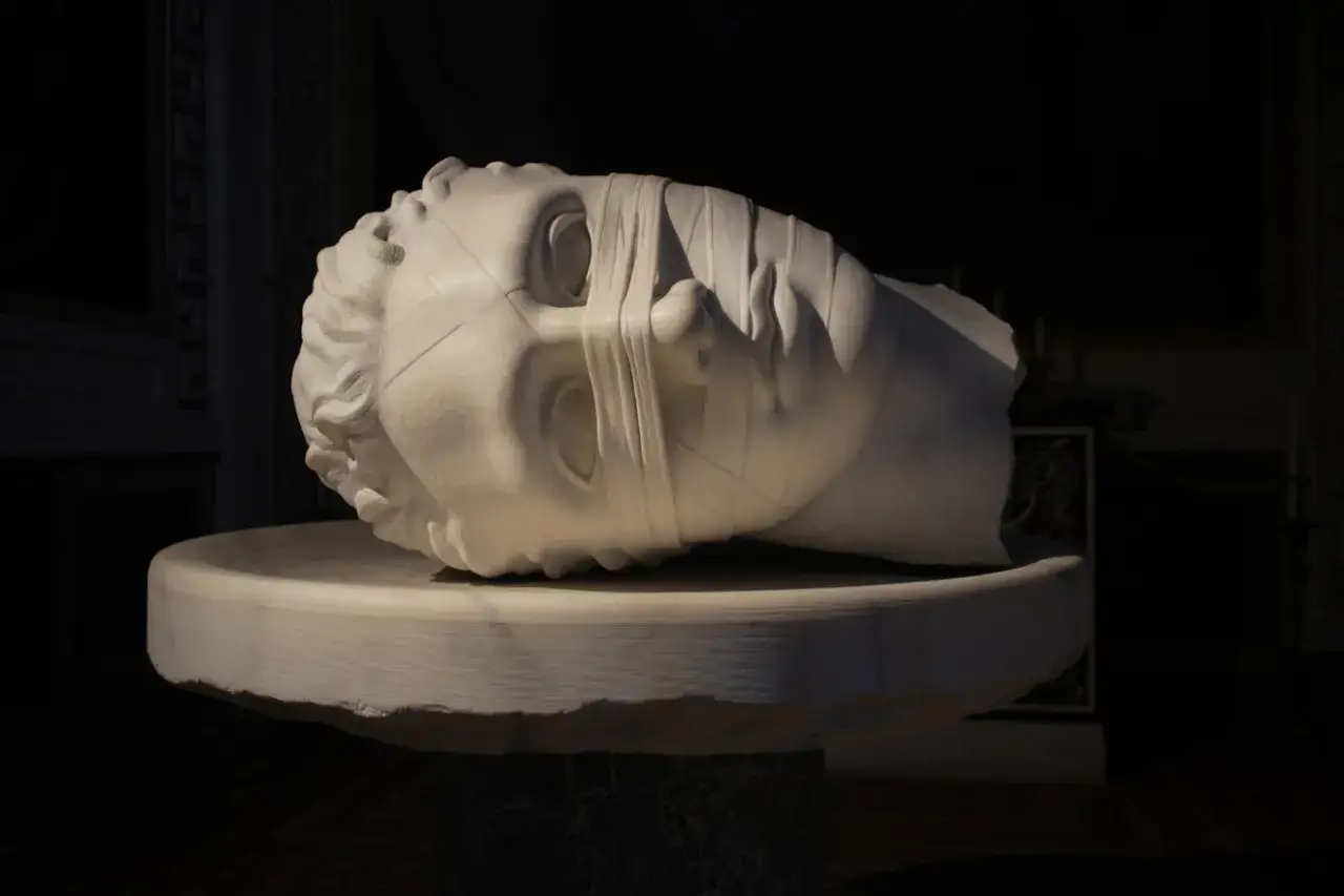 Biała marmurowa rzeźba głowy z opaską na oczach, leżąca na kamiennym postumencie.