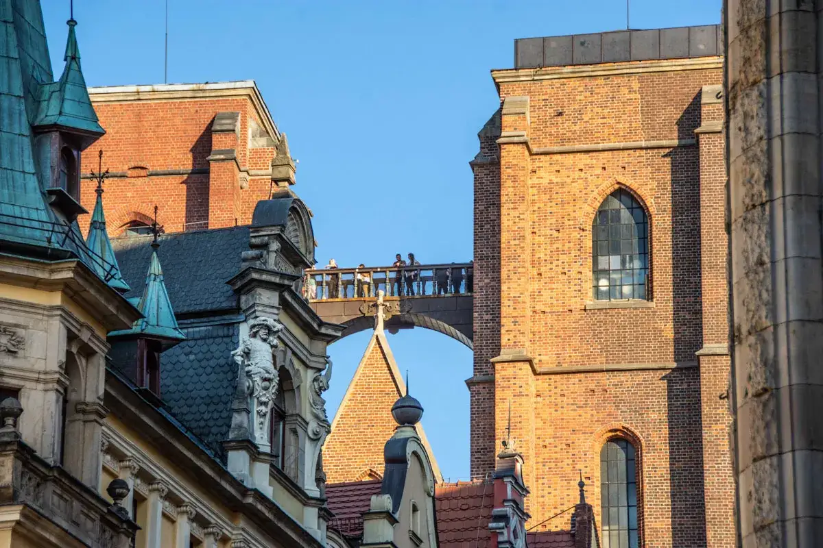 Most zabytkowych budynków we Wrocławiu z ludźmi na moście i wieżą Katedry w tle.