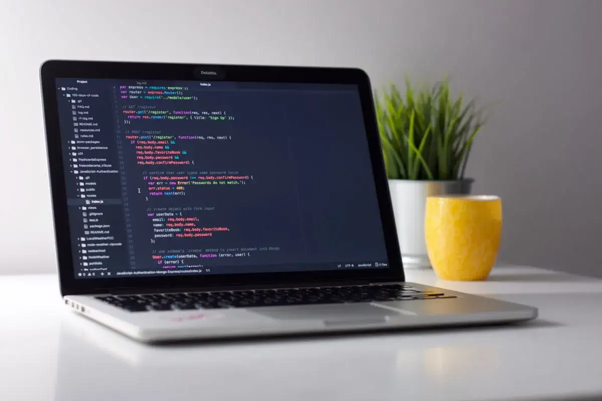 Laptop z kodem HTML na ekranie, obok zielona roślina i żółty kubek.