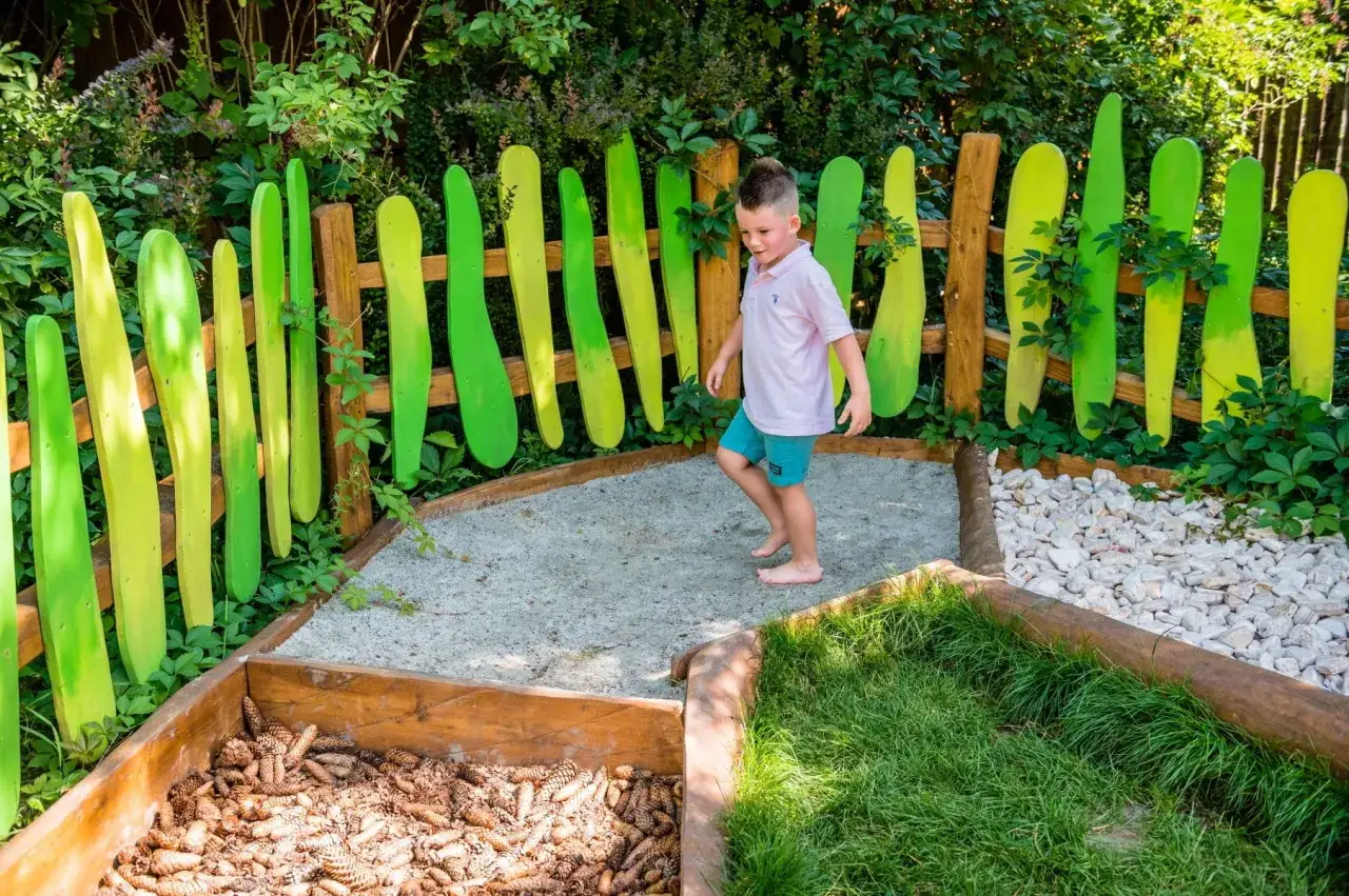 Chłopiec na ścieżce sensorycznej z piaskiem, szyszkami i trawą, otoczonej kolorowym płotkiem.