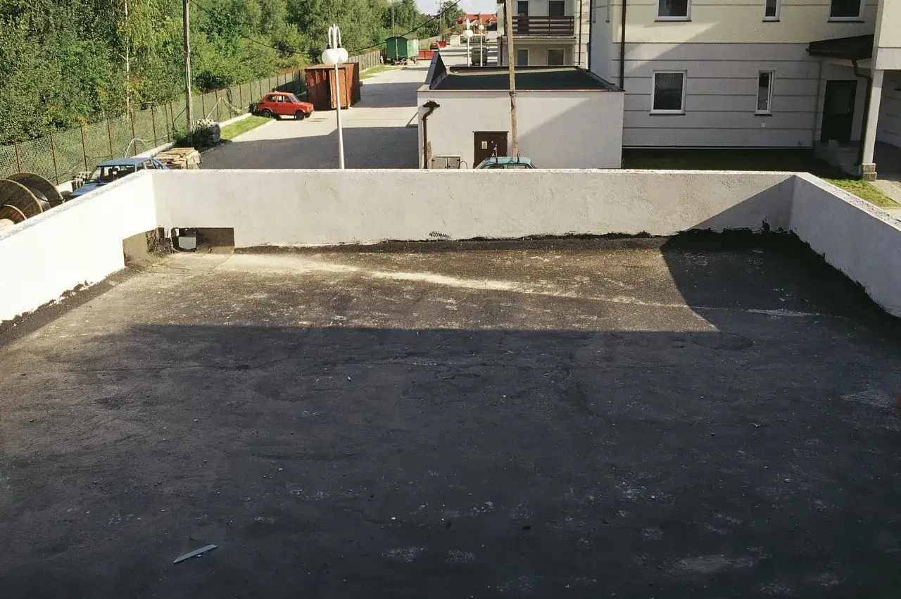 Płaski dach z widocznym systemem odwodnienia. W tle budynki i drzewa.