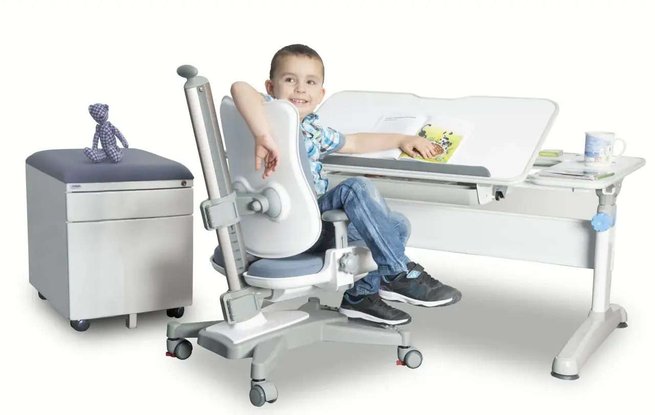 Uśmiechnięty chłopiec siedzi przy biurku na ergonomicznym krześle, czytając książkę.