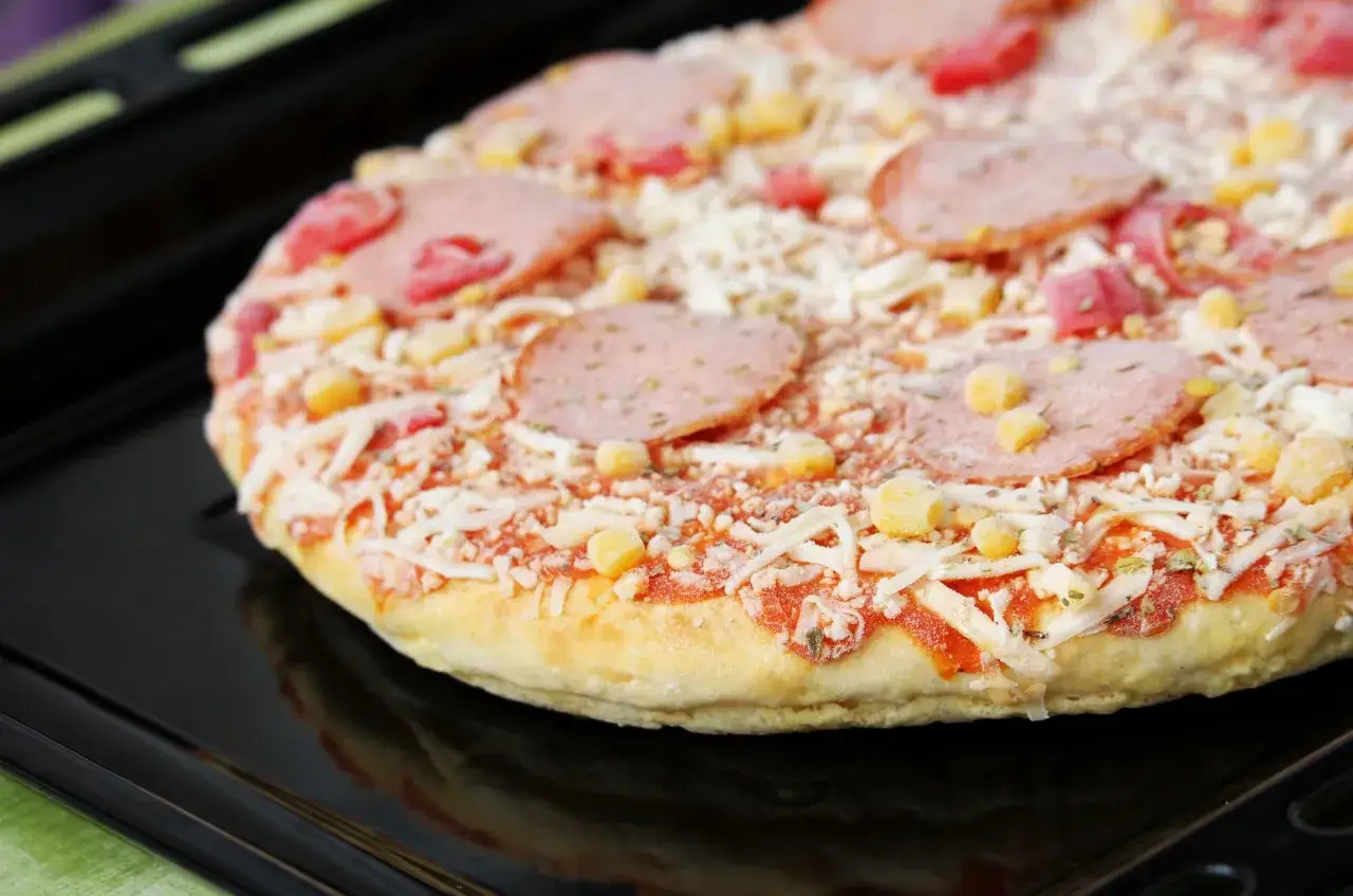 Mrożona pizza z serem, salami i kukurydzą na czarnej blasze do pieczenia.