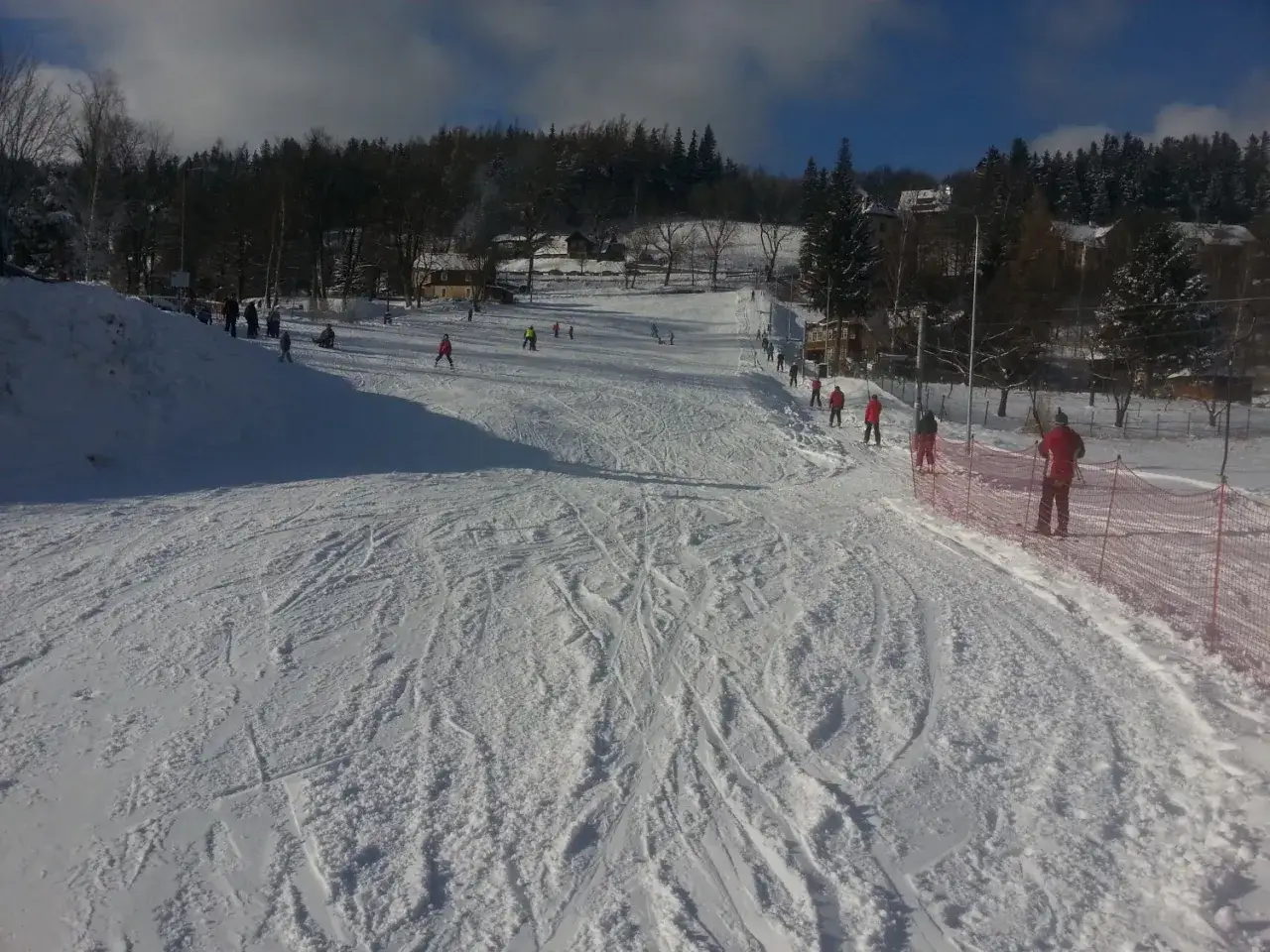 Stok narciarski w Karpaczu. Ludzie jeżdżą na nartach po ośnieżonym stoku, w tle las i budynki.