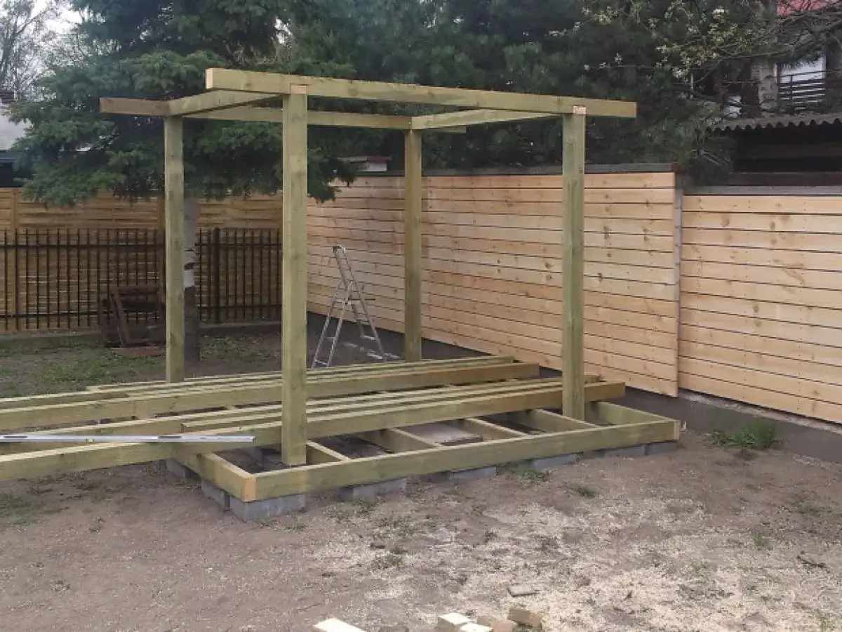 Konstrukcja altany w trakcie budowy. Drewniany szkielet i podłoga na bloczkach. W tle płot.