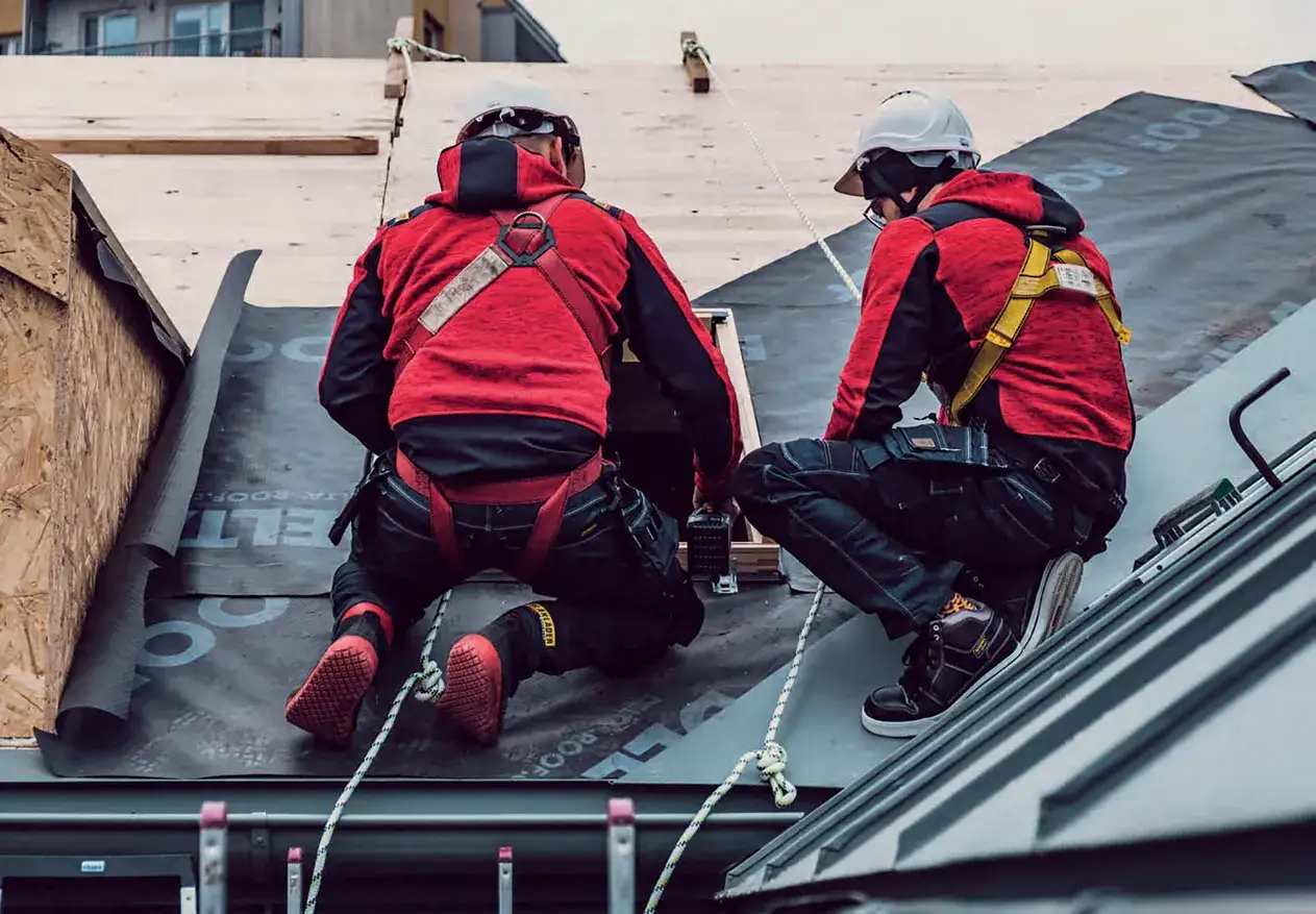 Dwóch dekarzy w czerwonych kurtkach i kaskach pracuje na dachu, zabezpieczonych linami.