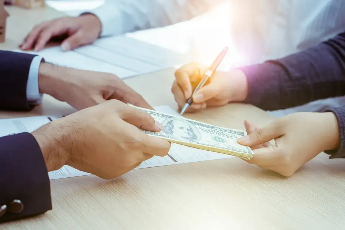 Dłonie przekazujące stos banknotów dolarowych, jedna osoba podpisuje dokument.