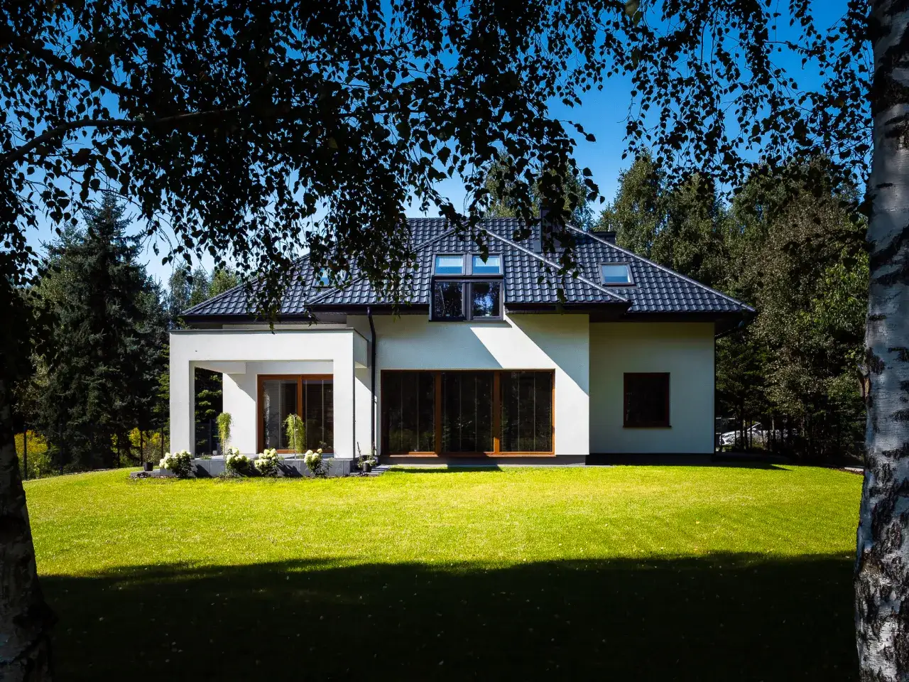 Nowoczesny, biały dom z dużymi oknami i zielonym trawnikiem, otoczony drzewami.