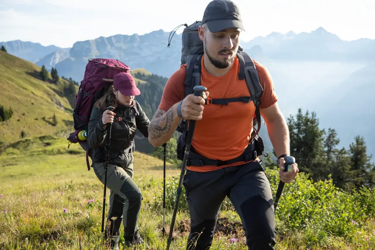 Dos excursionistas con mochilas y bastones caminan por una ladera montañosa.