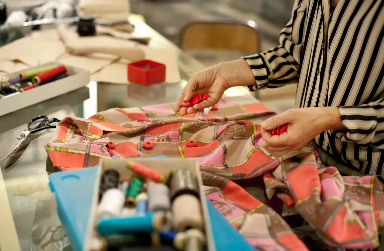 Ręce układają czerwone guziki na wzorzystej tkaninie. Widać pudełko z kolorowymi nićmi i nożyczki. To wszystko to pasmanteria, co to jest, można zobaczyć na tym zdjęciu.