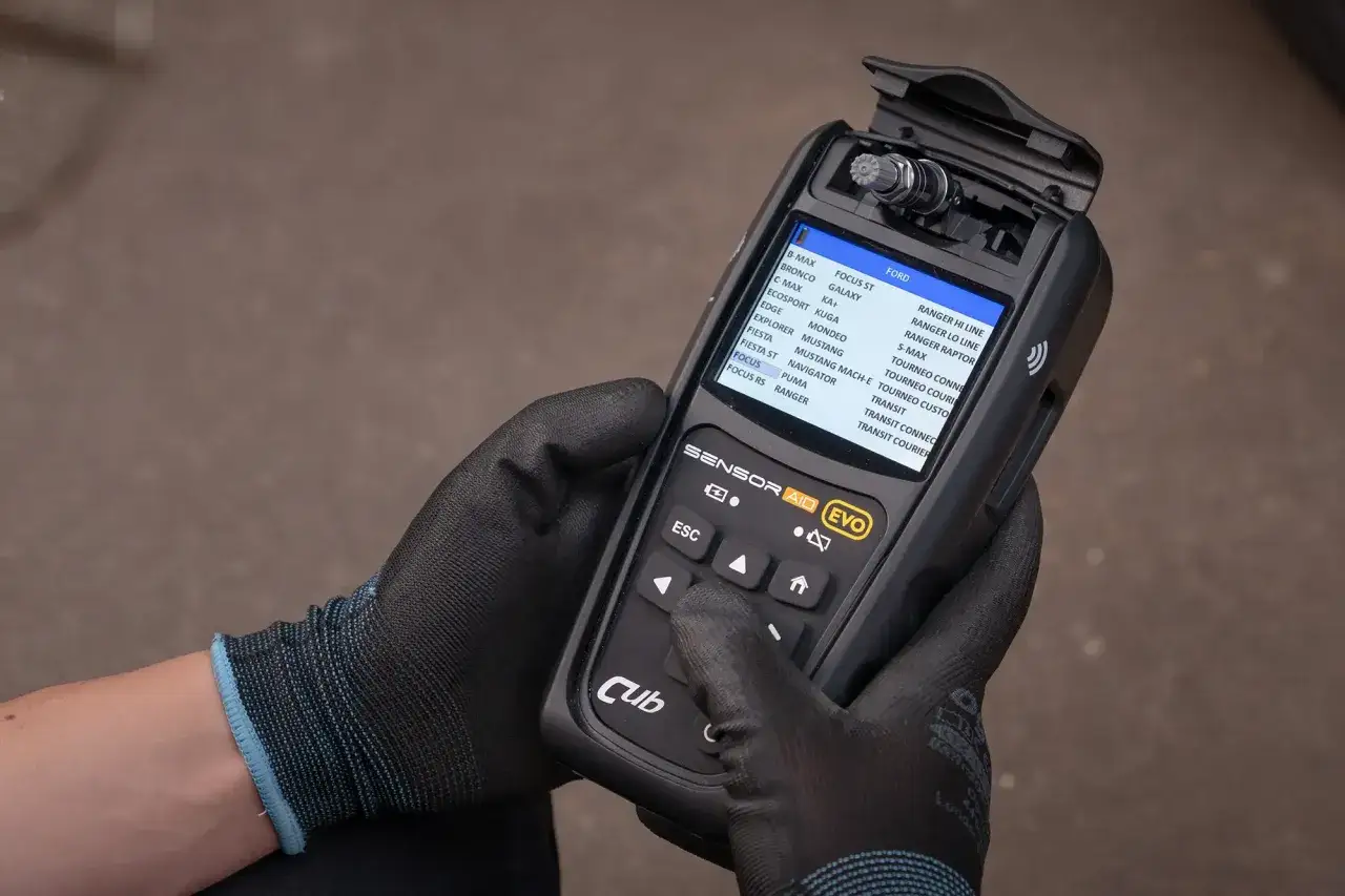 Ręce w rękawiczkach trzymają urządzenie diagnostyczne "Sensor Aid EVO" z listą modeli Forda. Zastanawiasz się, ile kosztuje czujnik?