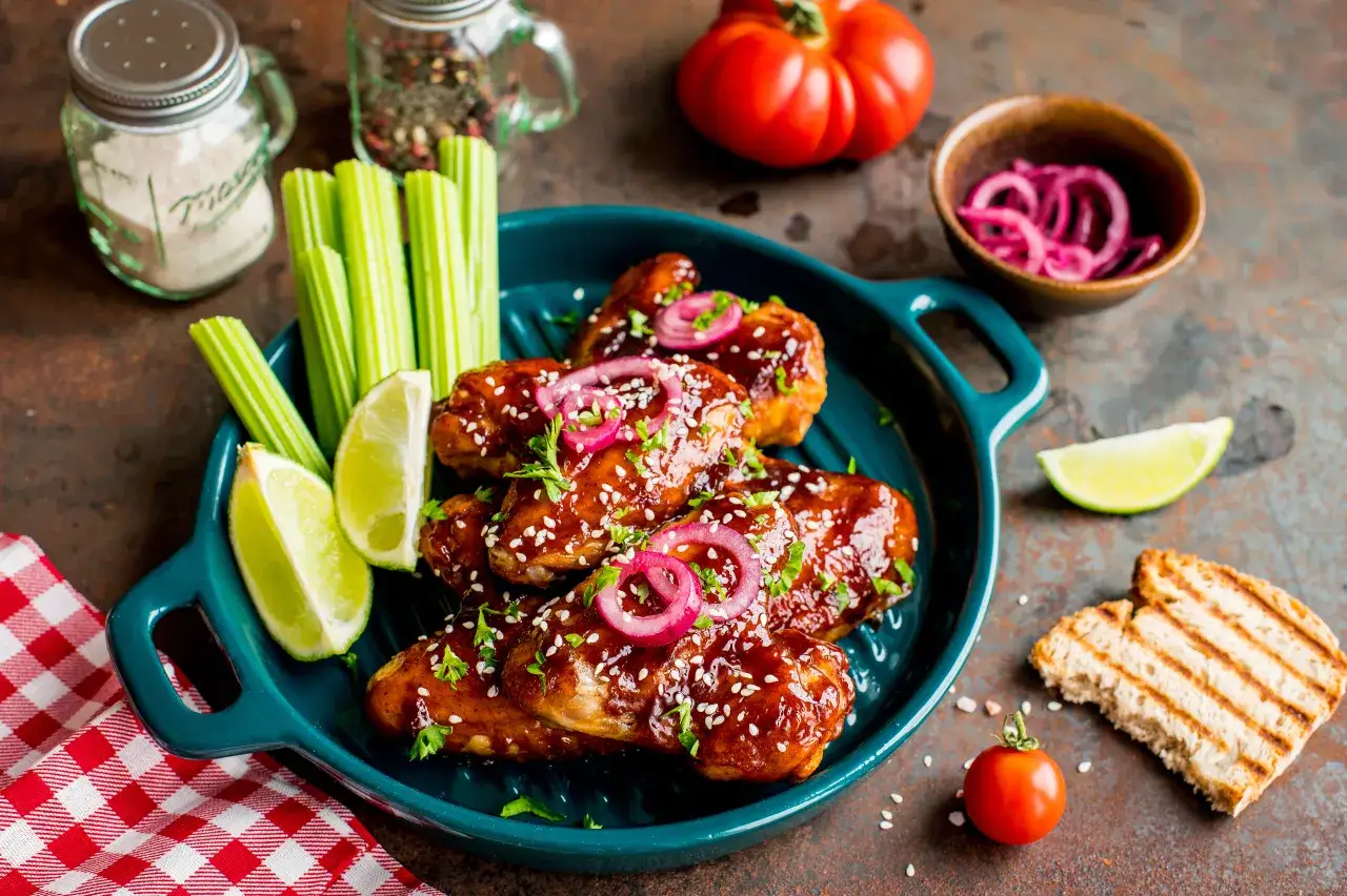 Skrzydełka z BBQ w sosie, podane z łódeczkami limonki, selerem i marynowaną cebulką. Obok grzanka i pomidorki.