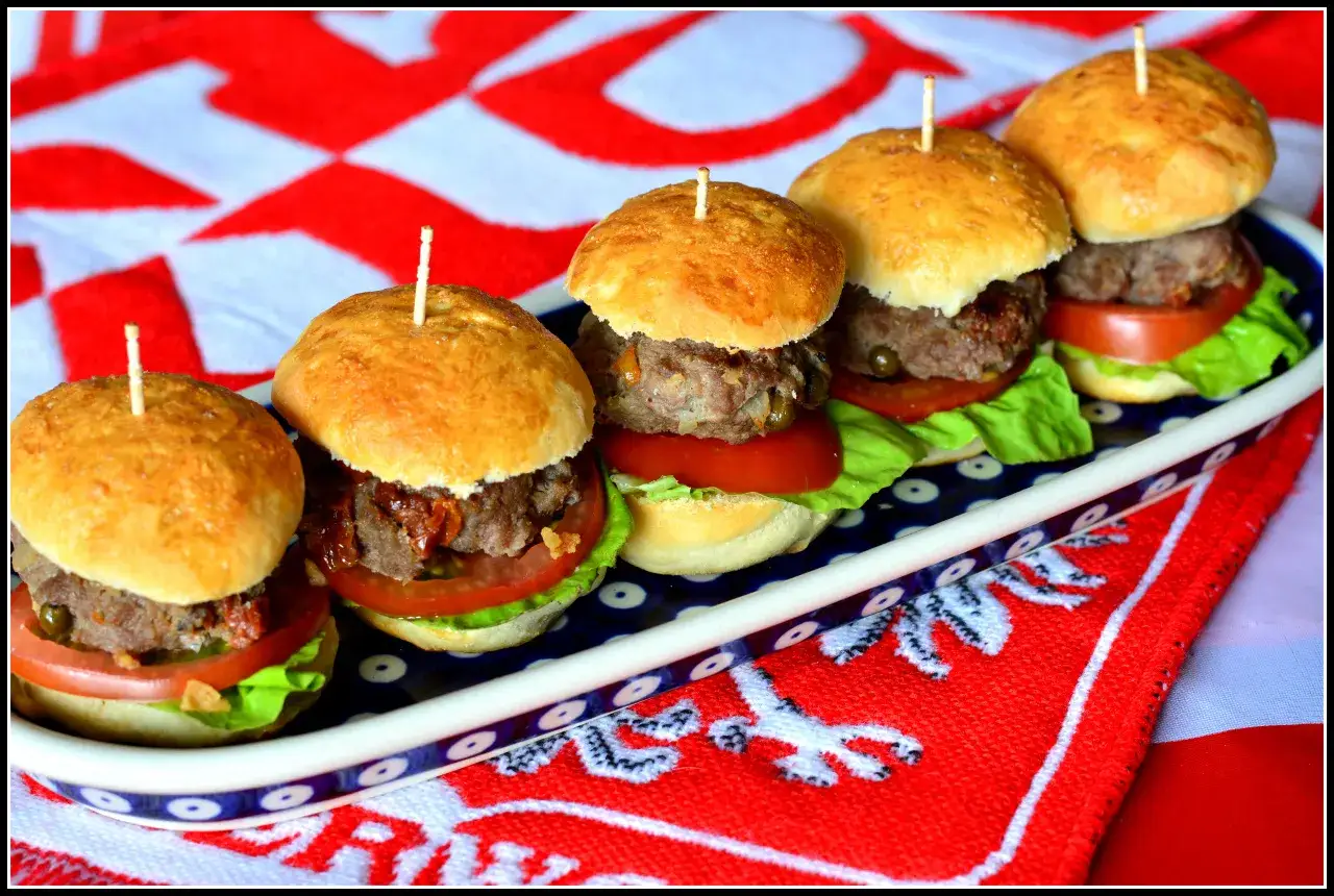 Rząd pięciu mini burgerów z mięsem, sałatą i pomidorem na talerzu.
