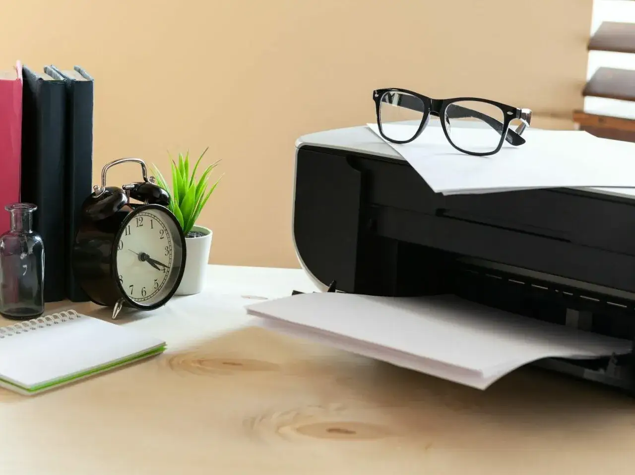 Drukarka z okularami na górze, obok budzik, książki i roślina.