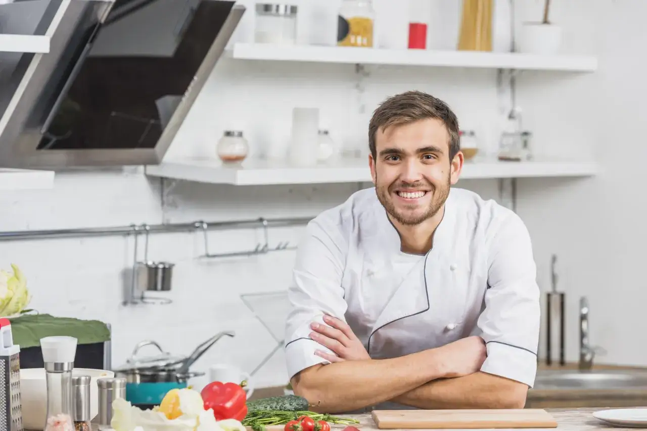 Uśmiechnięty kucharz w kuchni, gotowy do pokazania, jak reklamować firmę cateringową. Świeże warzywa na blacie.
