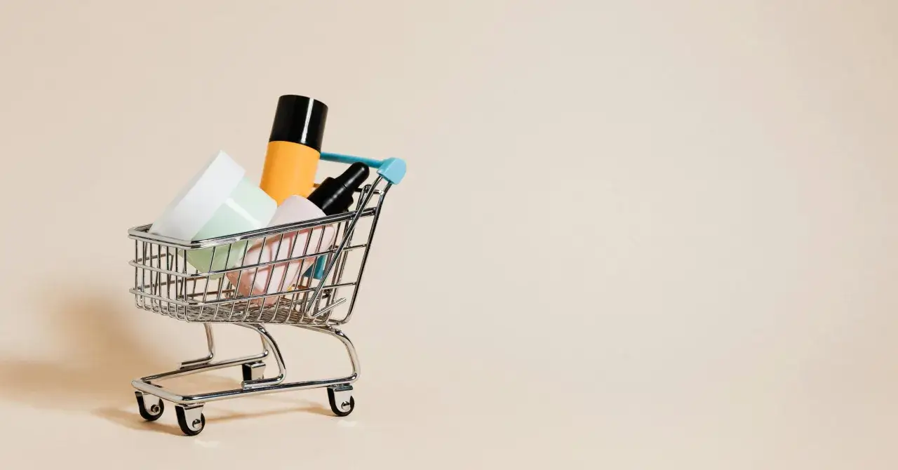 Miniaturowy wózek sklepowy wypełniony kosmetykami na jasnym tle.
