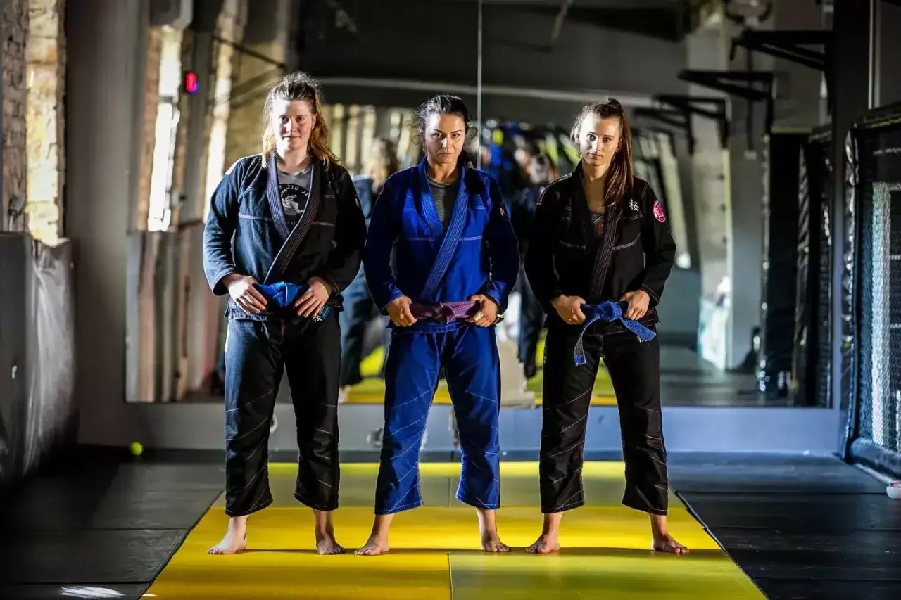 Trzy kobiety w kimonach do BJJ. Środkowa ma purpurowy pas bjj, a pozostałe niebieskie.