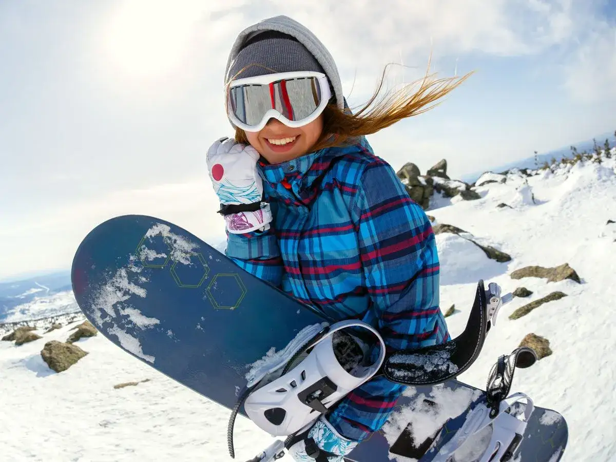 Uśmiechnięta snowboardzistka w goglach i czapce trzyma deskę snowboardową na tle zaśnieżonych gór.