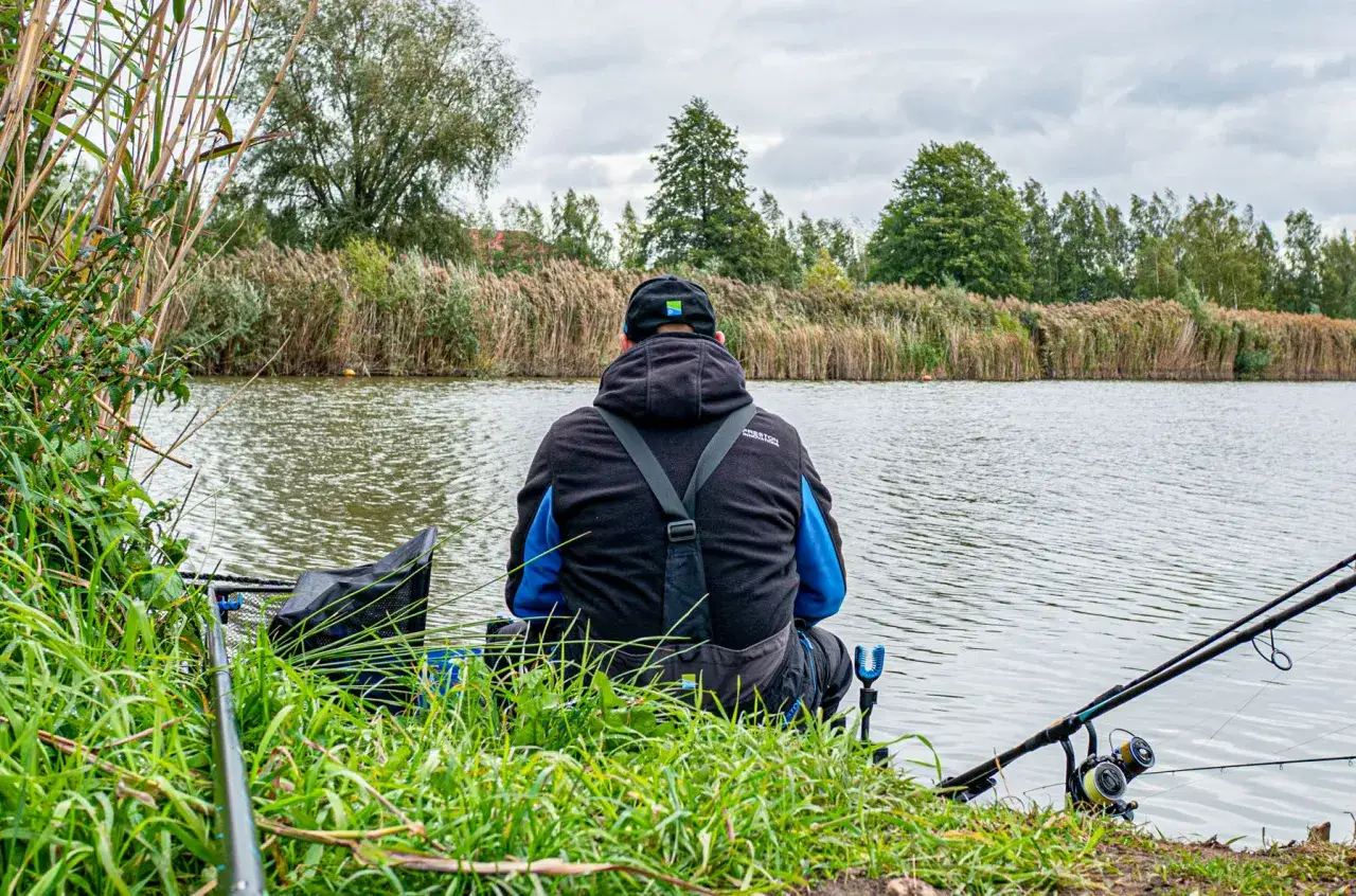 Wędkarz siedzi nad rzeką, przygotowując zanętę na rzekę feeder. W tle trzciny i drzewa.