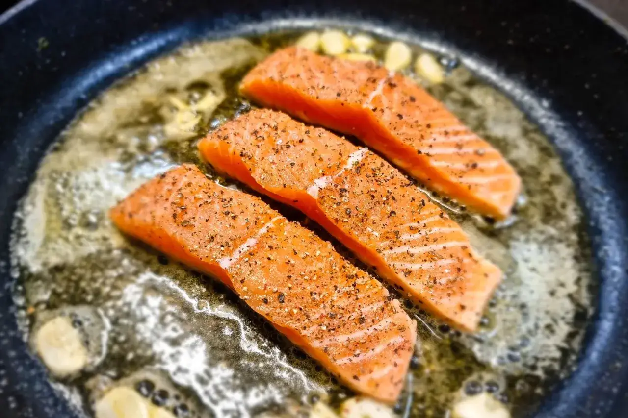Trzy kawałki łososia smażą się na patelni z masłem i czosnkiem. Idealny sposób, by dowiedzieć się, ile smażyć rybę na patelni.