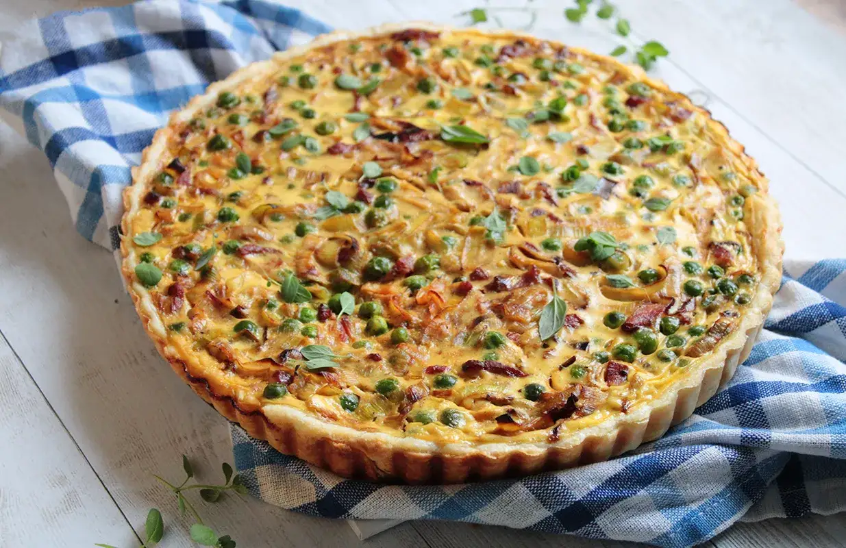 Pyszna tarta z porem i boczkiem, udekorowana groszkiem i ziołami, na niebiesko-białej kratce.
