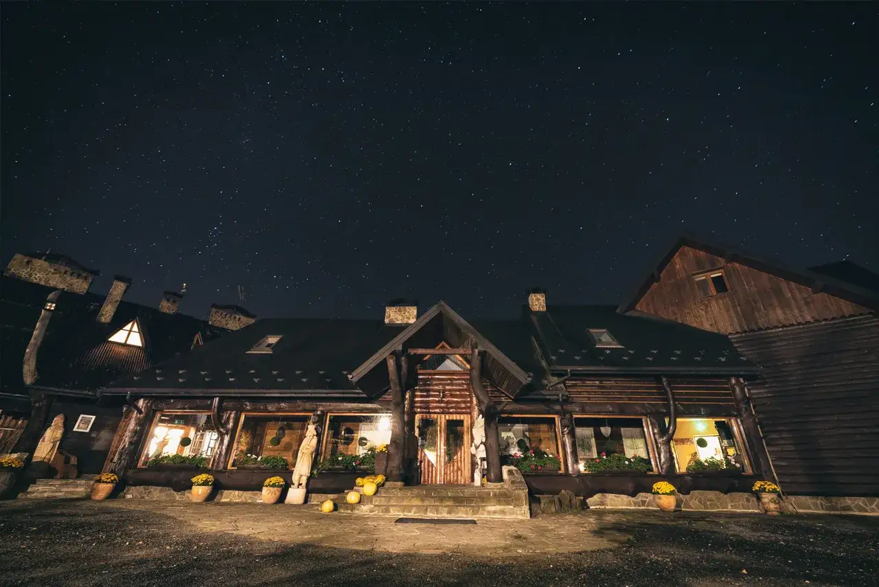 Nocne niebo nad bacówką Biały Jeleń. Drewniany budynek z oknami oświetlonymi ciepłym światłem, otoczony donicami z kwiatami.