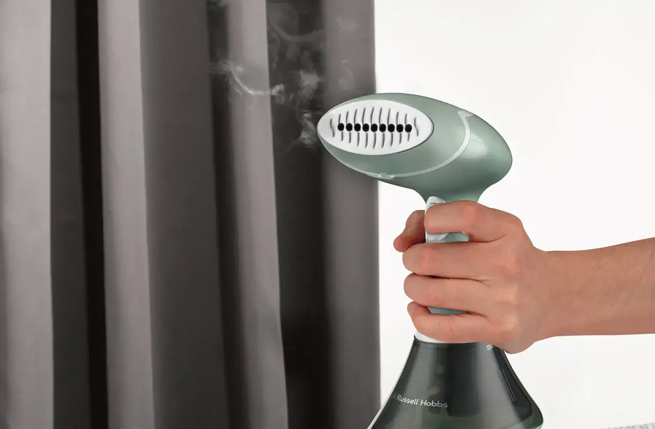 Dłoń trzyma parownicę Russell Hobbs, z której wydobywa się para, zbliżając ją do szarych zasłon.