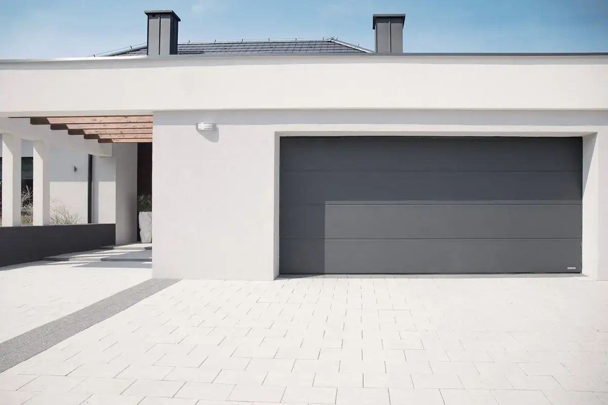 Nowoczesny dom z szarym, segmentowym, ciepły próg bramy garażowej. Podjazd z białej kostki brukowej.