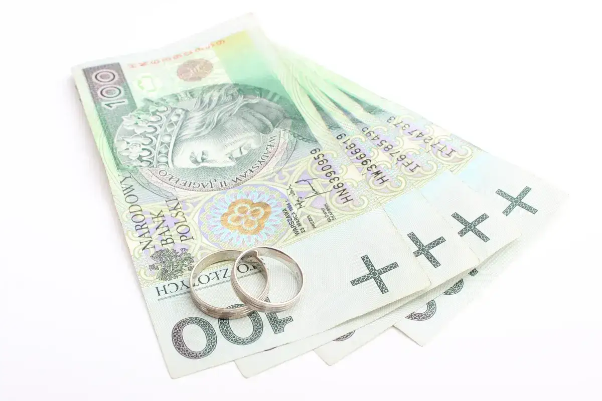 Dwa obrączki na stosie banknotów 100 zł. Symbolizuje to finansowe aspekty małżeństwa, co to jest intercyza.