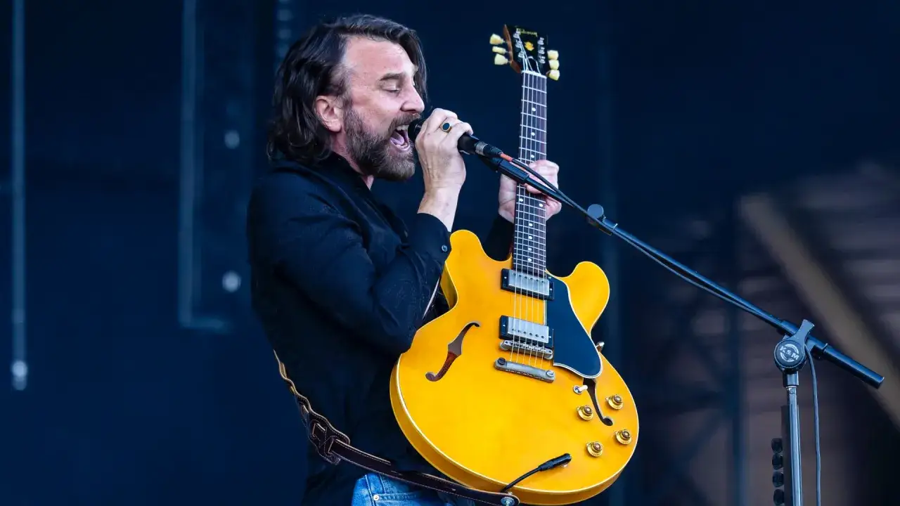 Mężczyzna z brodą i kręconymi włosami śpiewa do mikrofonu, trzymając żółtą gitarę. Czuje, że "kto śpiewa mój jest ten kawałek podłogi".