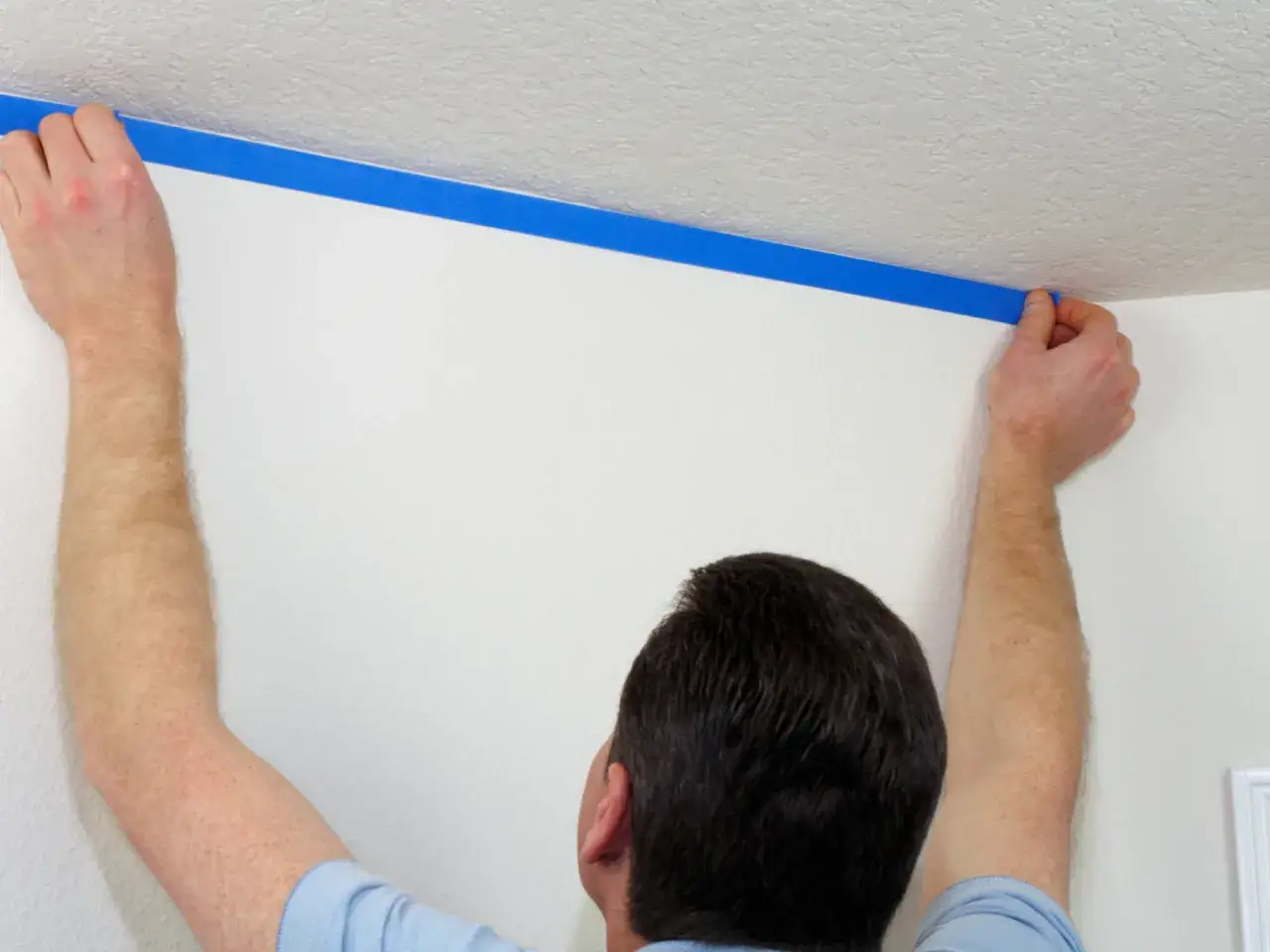 Mężczyzna przykleja niebieski pasek pod sufitem, przygotowując ścianę do malowania.