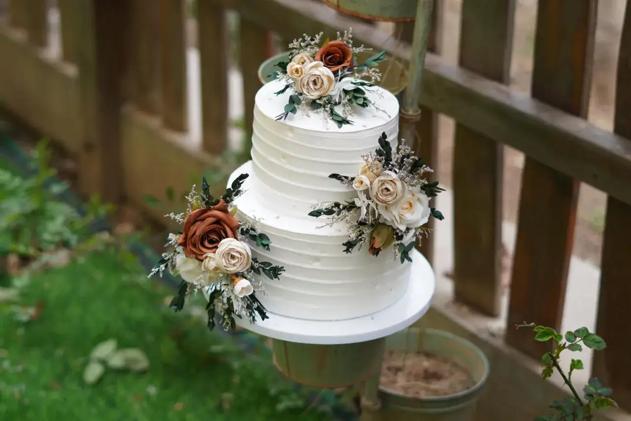 Elegancki tort weselny z białym kremem, ozdobiony bukietami kwiatów w odcieniach brązu i kremu. Wygląda jak tort z margaretkami, idealny na rustykalne przyjęcie.