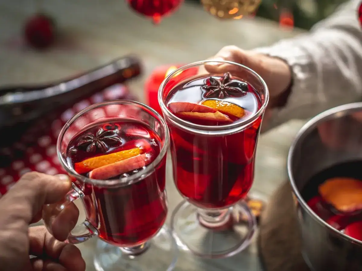 Dwa kieliszki grzanego wina z pomarańczą i anyżem, w tle butelka i garnek.