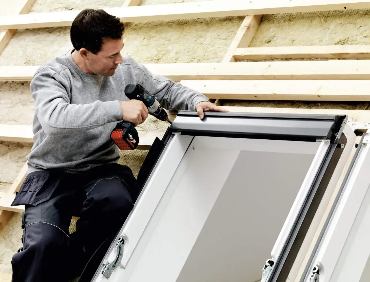 Fachowiec montuje okna dachowe, używając wkrętarki. Prace na poddaszu postępują.