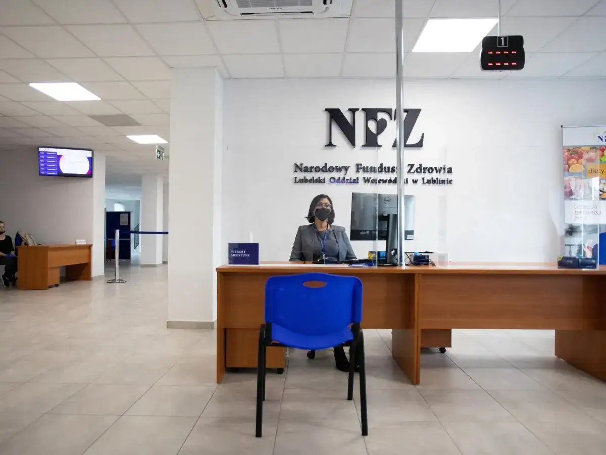 Pracownica NFZ za ladą w biurze, z logo NFZ na ścianie.