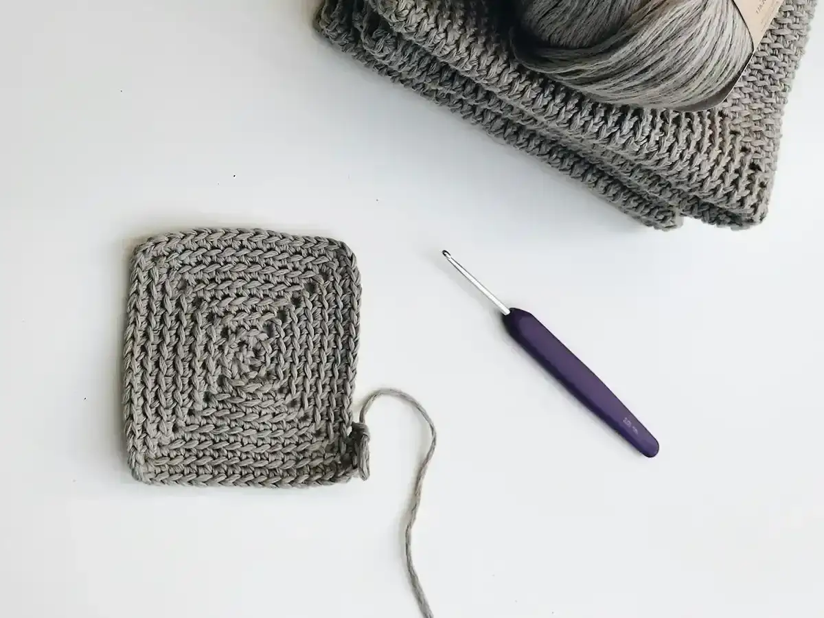 Szary kwadrat robiony na szydełku, włóczka i fioletowy szydełko. Idealne do nauki szydełkowania krok po kroku.