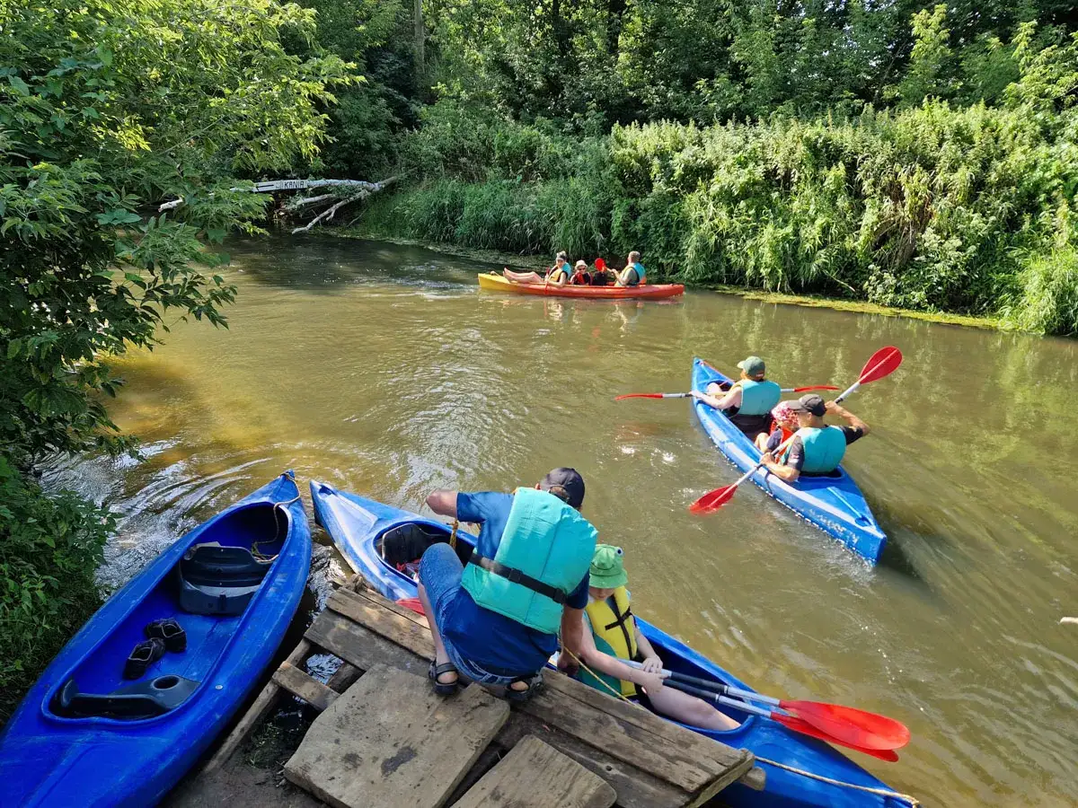 Rodzina w kajakach na rzece, przygotowująca się do spływów kajakowych Prosną. Zielone brzegi i słoneczny dzień.