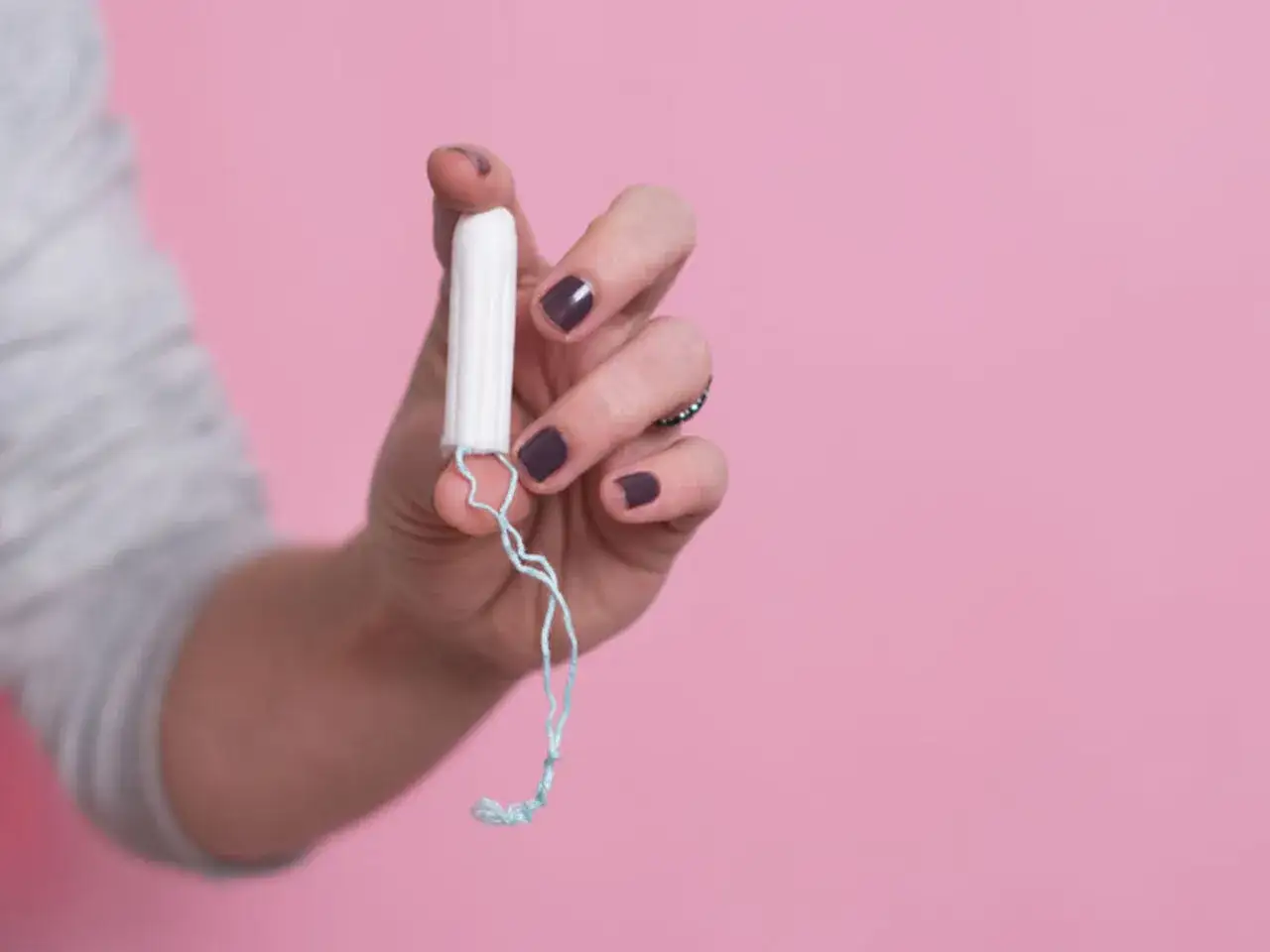 Dłoń trzymająca biały tampon z niebieskim sznurkiem na różowym tle.