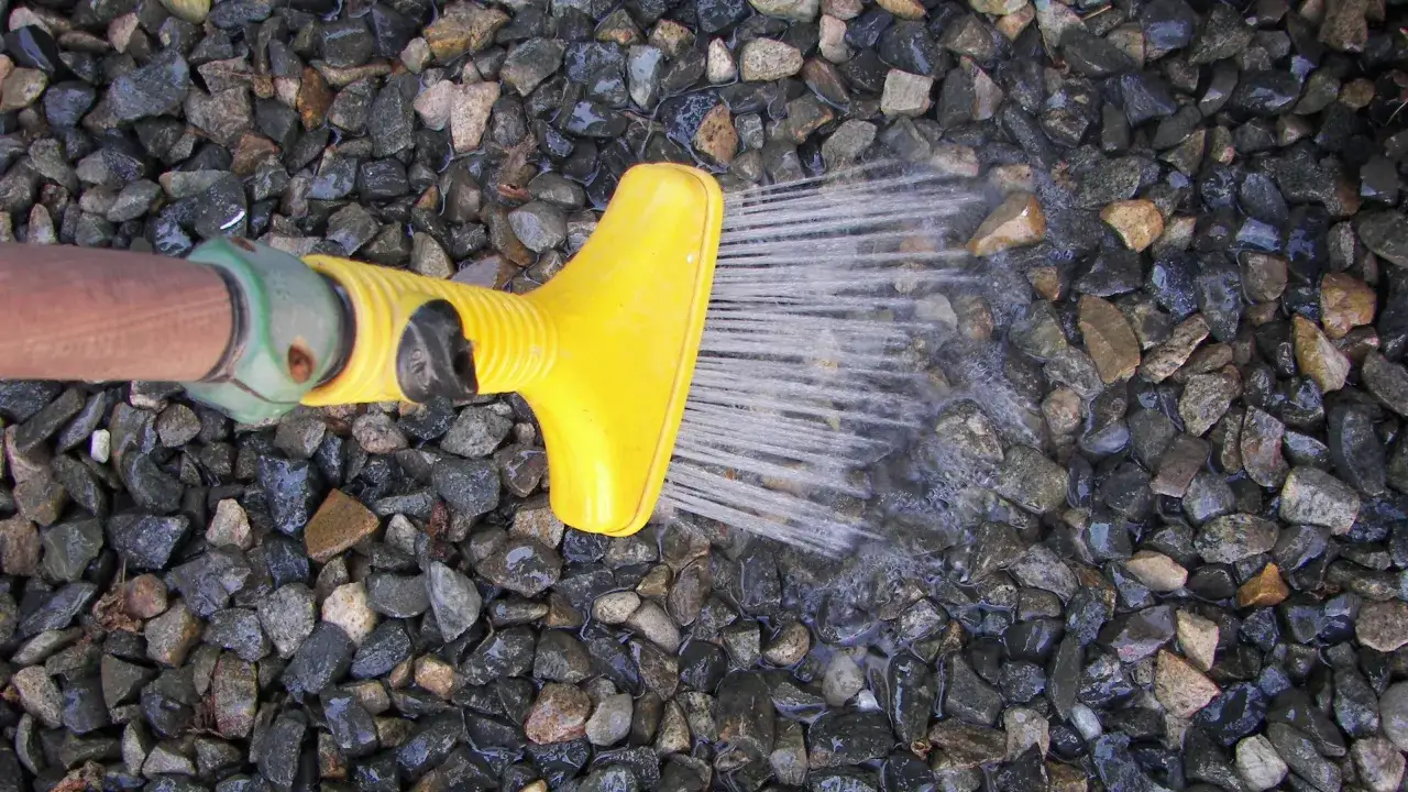 Żółta końcówka węża ogrodowego spryskuje wodą kamyczki, pokazując, jak wyczyścić kamyczki w ogrodzie.