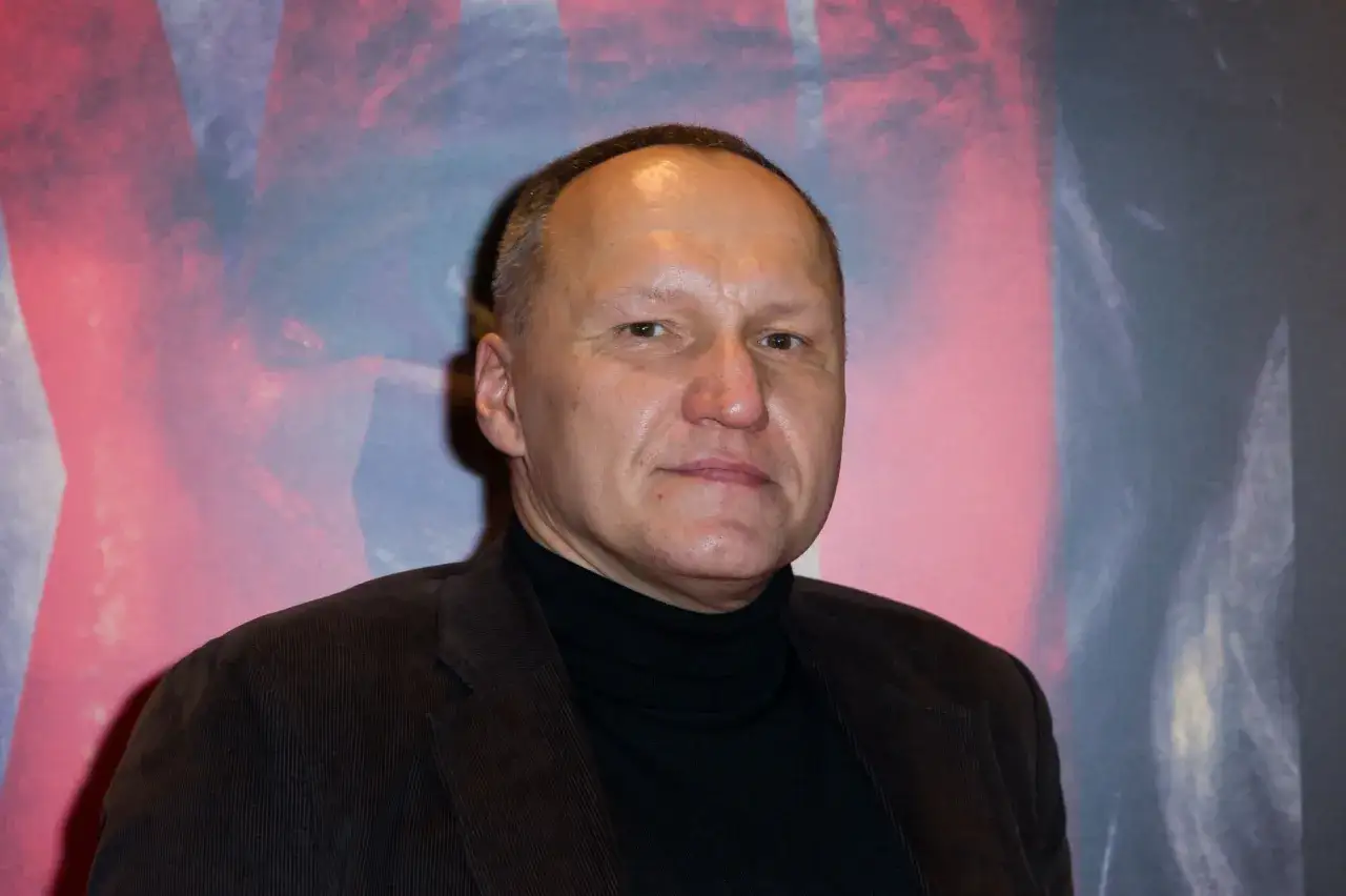 Portret Mariusza Grzegorzka w czarnym golfie i brązowej marynarce na tle abstrakcyjnego wzoru.