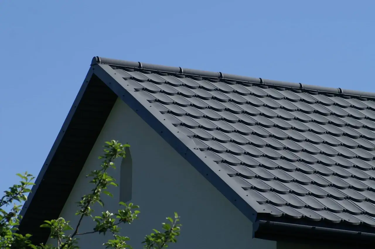 Szary dach z dachówki ceramicznej pod błękitnym niebem, z fragmentem ściany domu i gałęziami drzew.
