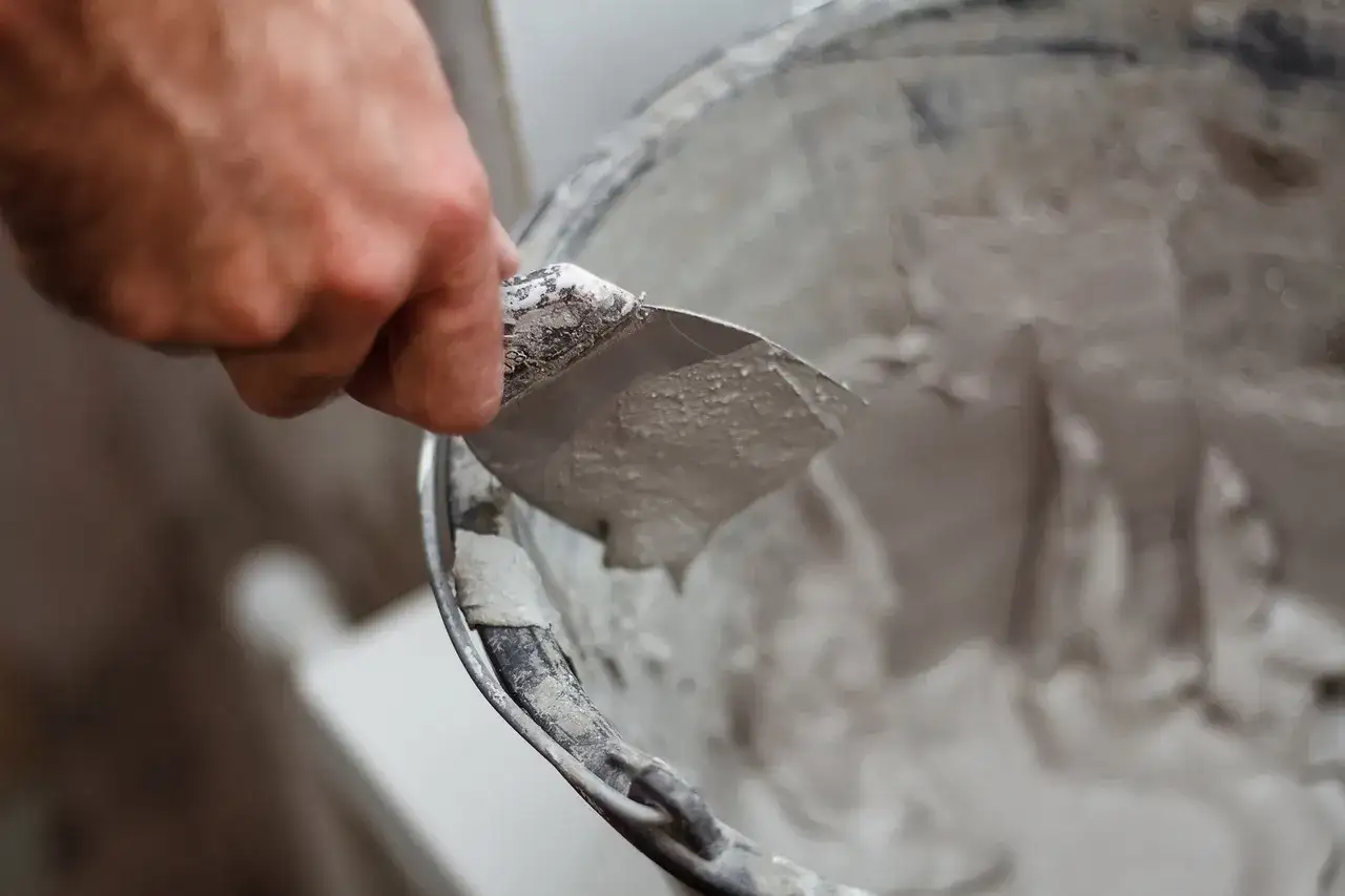 Dłoń trzymająca kielnię z gipsową zaprawą nad wiadrem.